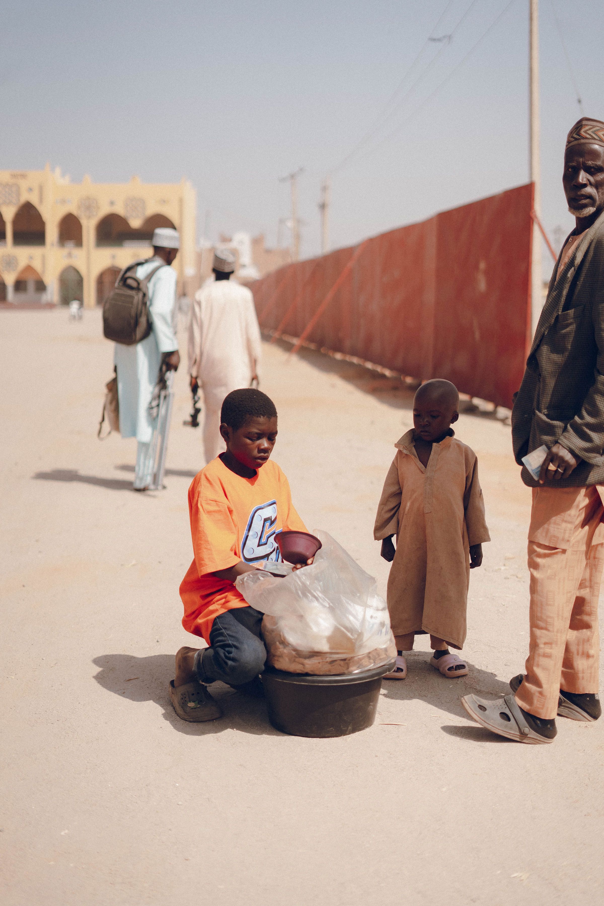 A boy sits with a bucket on a dusty street while two men and a child stand nearby